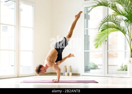 Muscular young man doing Flying Crow yoga pose, working out indoors against bright windows and white wall Stock Photo