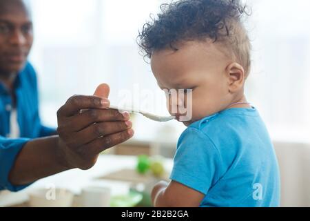 Side view portrait of cute African-American toddler refusing to eat food from spoon in kitchen interior, copy space Stock Photo