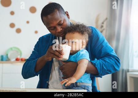 Waist up portrait of happy African-American father giving drink to cute little boy in kitchen interior, copy space Stock Photo