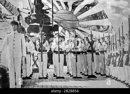 The crew of a ship of the Imperial Japanese Navy lined up on the deck. In the background a large flag of the Navy is waving. (Undated photo, approx. 1920s) Stock Photo