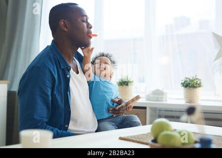 Portrait of happy African father playing with little son while sitting in sunlit kitchen, copy space Stock Photo