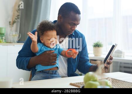 Side view portrait of African-American man holding crying baby while using digital tablet in home interior, copy space Stock Photo