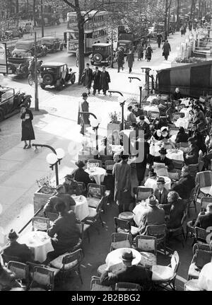People sit on the terrace of Cafe Kranzler on the Kurfuerstendamm in Berlin. The outside area of the cafe is bordered by flower troughs, which are illuminated. On the left there is a double-decker bus, typical for Berlin. Stock Photo