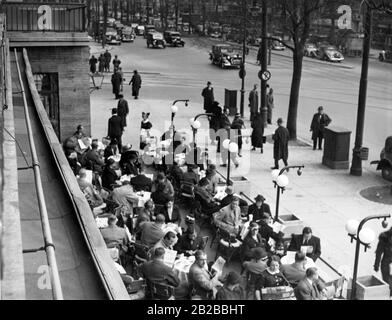 Guests sit on the terrace of Cafe Kranzler on the Kurfuerstendamm in Berlin. On the right passers-by walk on the sidewalk. Stock Photo