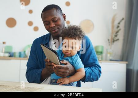 Portrait of African-American man holding crying baby while using digital tablet in home interior, copy space Stock Photo