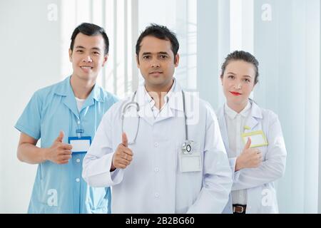 Group of happy multi-ethnic medical workers in scrubs showing thumbs-up and smiling at camera Stock Photo
