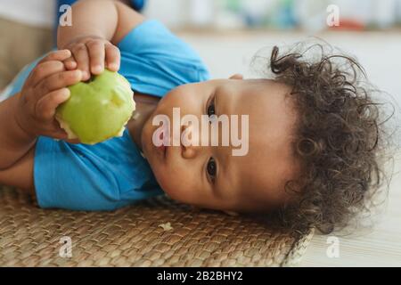 Portrait of cute mixed race toddler eating green apple while lying on changing table, copy space Stock Photo