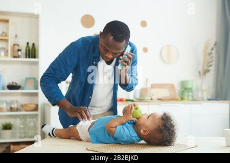 Portrait of mature African man calling wife while changing diaper to baby in home interior, copy space Stock Photo