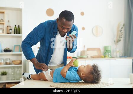 Portrait of mature African-American man calling wife while changing diaper to baby in home interior, copy space Stock Photo