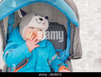 little boy sitting in the stroller in the winter and eats cake Stock Photo