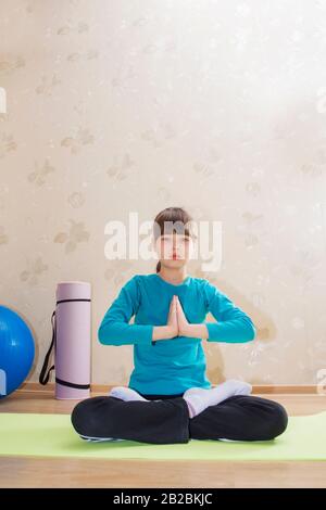 Young beautiful athletic girl practicing indoor yoga sitting in Ardha Padmasana with palms in Namaste, Half Lotus Posture. Stock Photo