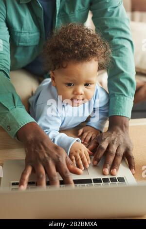 Vertical portrait of cute African-American baby using laptop and looking at screen with father, copy space Stock Photo