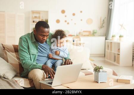 Portrait of mature African-American man working on laptop while babysitting son at home, copy space Stock Photo