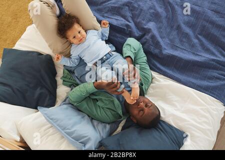Above view portrait of happy African-American man playing with cute baby son while lying on bed at home, copy space Stock Photo