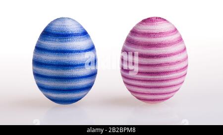 Two standing Easter eggs striped in male blue and female pink color on white background. Colored streaked egg shells wrapped by glued thin cotton yarn. Stock Photo