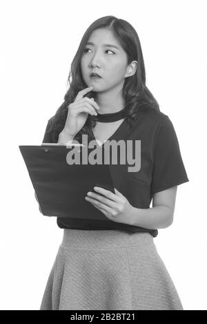 Young beautiful Asian businesswoman holding clipboard while thinking Stock Photo