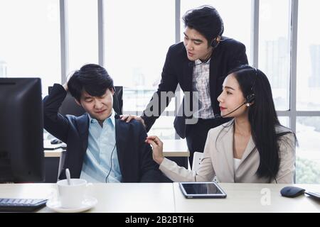 Two Asian call center agents cheer their colleague up who has problem about his work, project or personal life. Stock Photo