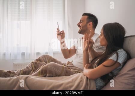 Young happy couple is looking at smartphone in bed in the morning. Stock Photo