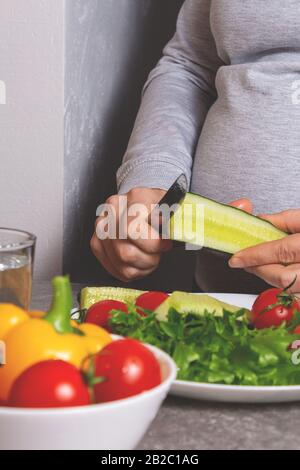 Unrecognizable pregnant woman cutting cucumber for salad in the kitchen. Cucumber, cherry tomatoes and fresh salad are already on the white plate. Hea Stock Photo