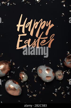 Happy Easter minimalistic concept with hand drawn modern lettering. Gilded eggs and shiny confetti on a black background. Stock Photo