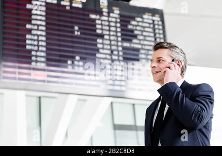 Attractive mature businessman talking on smartphone while standing against flight screen display in airport Stock Photo