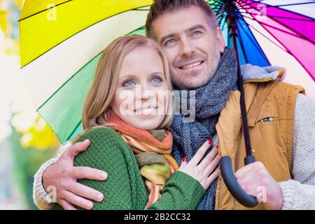 Portrait of attractive man holding umbrella while embracing his wife in park Stock Photo