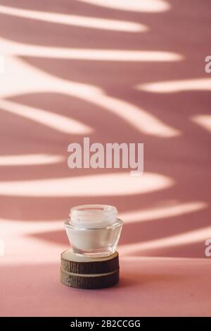 Glass jar with white moisturizing cream on wooden saw cuts on a pink background. Lotion on the background of the tropical shadow of a palm leaf. The c Stock Photo