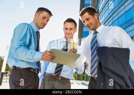 Mature attractive businessman showing and discussing plans over digital tablet with colleagues against office building