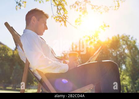 Portrait of handsome businessman drinking coffee and reading newspaper while sitting on folding chair in park Stock Photo