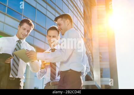 Mature attractive businessman showing and discussing plans over digital tablet with colleagues against office building Stock Photo