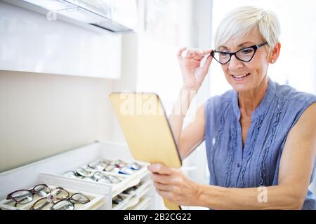 Beautiful mature woman choosing new pair of spectacles in opticians store. Eyesight correction. Stock Photo