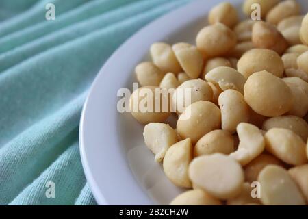 Close up side view of roasted Macadamia Nuts on a white plate with a pale mint green table cloth background, with copyspace to left. Stock Photo