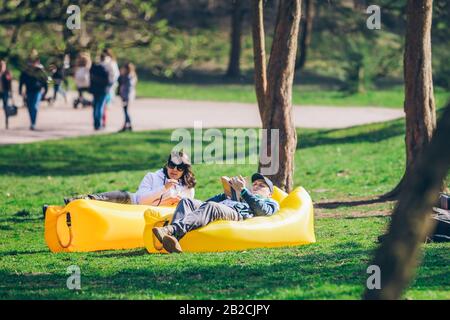 couple laying on yellow inflatable mattress in city park. reading book. drinking smoothies Stock Photo