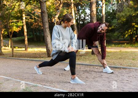 Photo of beautiful sporty couple in sportswear doing exercises while working out in green park during sunny summer day Stock Photo