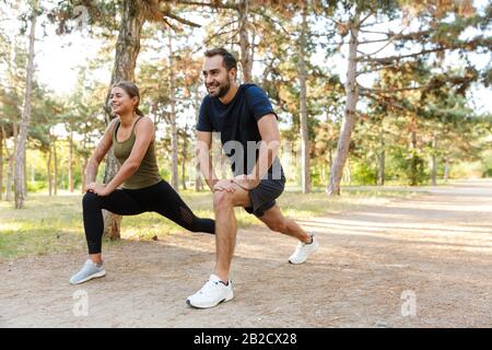 Photo of attractive smiling couple in sportswear doing exercises while working out in green park during sunny summer day Stock Photo