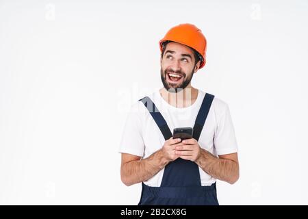 Image of a cheerful young man builder in helmet posing isolated over white wall background using mobile phone. Stock Photo