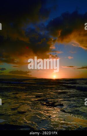 Dramatic sunset with rain showers falling from storm clouds over ocean, reflection in volcanic rocks, on the North Shore of Oahu, Haleiwa, Hawaii, USA Stock Photo