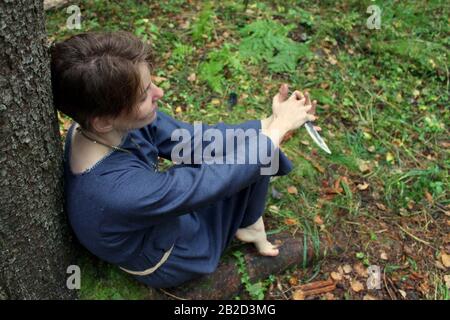 Syktyvkar, Russia - september, 2019: a woman of european appearance with short dark hair, dressed in a blue robe, performs a pagan witchcraft ritual. Dark forest on background. Stock photography. Stock Photo