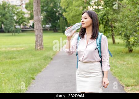 body hydration. woman in casual clothes and backpack drinking water from a bottle in park Stock Photo