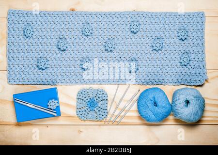 On background of wooden table is fragment of crocheted openwork napkin with decorative elements, knitting tools, blue balls of yarn close up. Backdrop Stock Photo