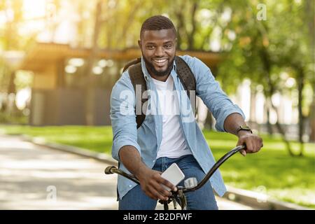 Handsome Black Man With Backpack And Smartphone Riding Bicycle, Posing Outdoors Stock Photo