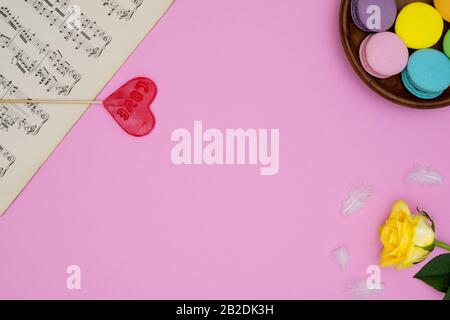 Pink background with antique music sheets, french macarons sweets, yellow rose, heart shaped lollipop love and small white soft feathers.Top view. For Stock Photo