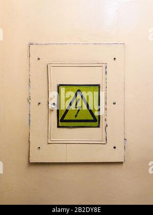 Locked electrical panel with attention sign on the wall painted in yellow oil paint. There is a high voltage sign on the door. Stock Photo