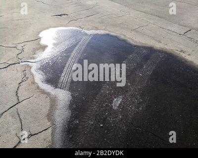 Track from a studded tire on the ice of a frozen puddle on cracked asphalt in the bright sunshine on a winter day. Stock Photo