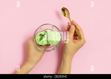 Woman hands holding glass cup with homemade avocado ice cream and golden spoon on light pink background. Top view, flat lay Stock Photo