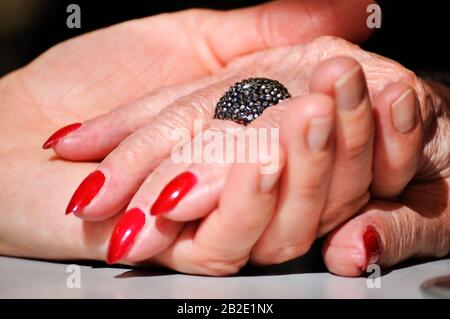 Adult daughter holding mother wrinkled old hand wearing a ring and red nail polish. Scene partially lit with a dramatic soft effect. Hands on the tabl Stock Photo