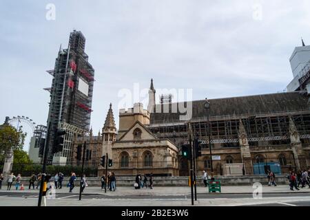 Tourists walk by Westminster Palace while Big Ben is covered with scaffolding while under restoration.  The London Eye can be seen in the background. Stock Photo