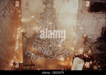 picture with expressions and words about Christmas wrapped in yellow garlands on a background of a wall and candles nearby. Stock Photo