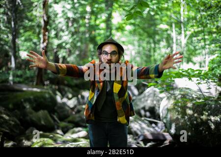 A funny bearded man in the forest. The funny facial expression of the young male in a multi-colored jacket and hat. A bohemian guy in the forest Stock Photo