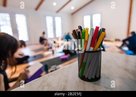 The small basket with multi-colored pencils. A little group of people taking a part in the yoga class at the background. The multi-colored handles Stock Photo